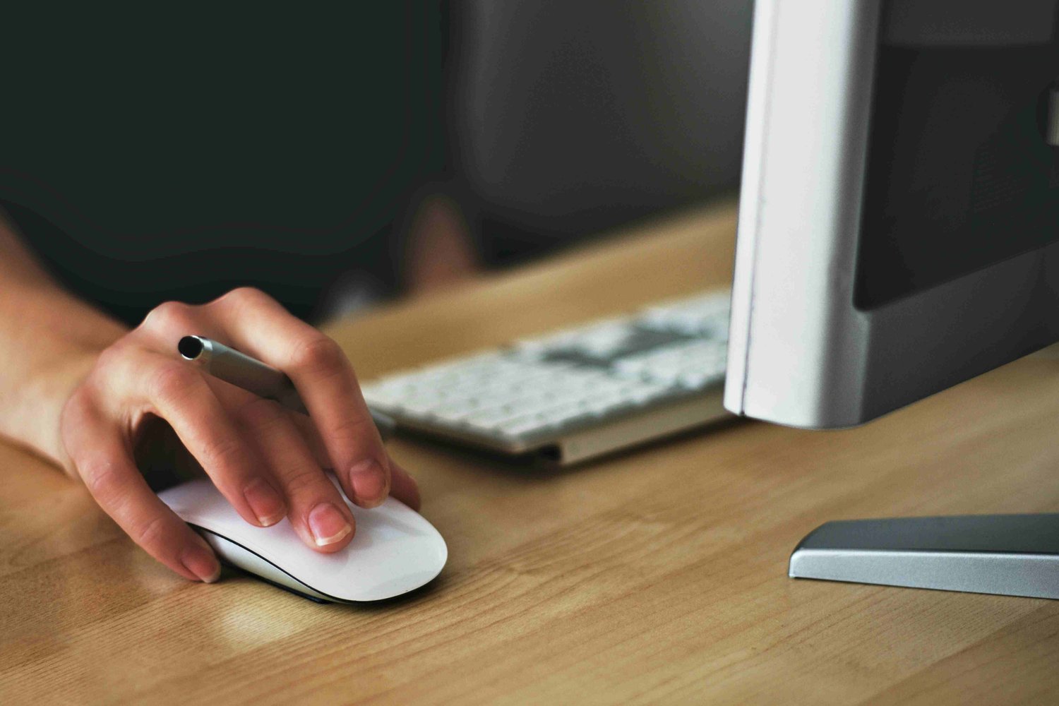 A close up view of a person's hand on a mouse, also holding a pen, sat at a desk. There is a keyboard and a monitor in front of them. 