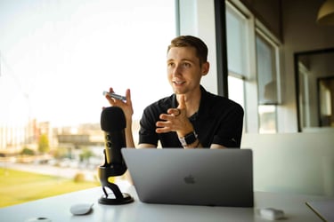 A young man with short hair sits at a desk, facing the camera. He's wearing a black t-shirt and a smart watch and is gesturing while speaking. In front of him on the desk is a Macbook and a USB microphone.