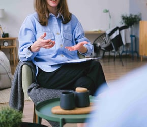 A person sits on a comfy chair in an office setting. They are gesturing with their hands, having a discussion with someone out of view. In front of them is a round, green coffee table with two blue mugs on top.